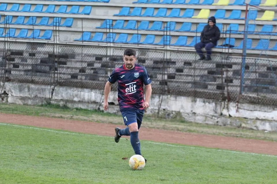 Un bărbat în echipament sportiv închis la culoare aleargă lângă o minge pe gazon; în spate se văd tribune cu scaune albastre.