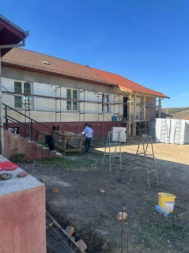 O clădire cu acoperiș de țiglă are schele în fațadă; două persoane se află în curte, unde sunt împrăștiate materiale și un recipient galben.