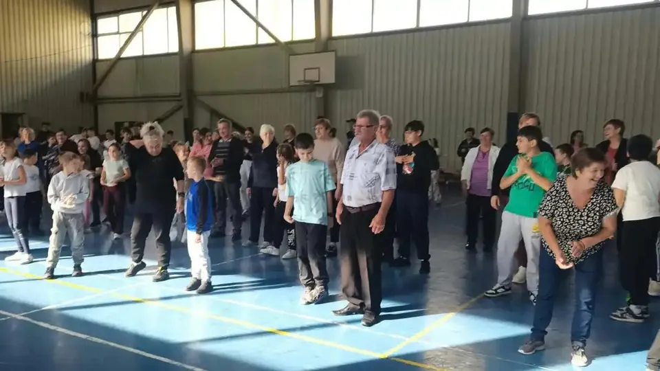 Mai multe persoane de vârste diferite stau în rânduri într-o sală de sport, purtând haine casual; în fundal se vede un panou de baschet.