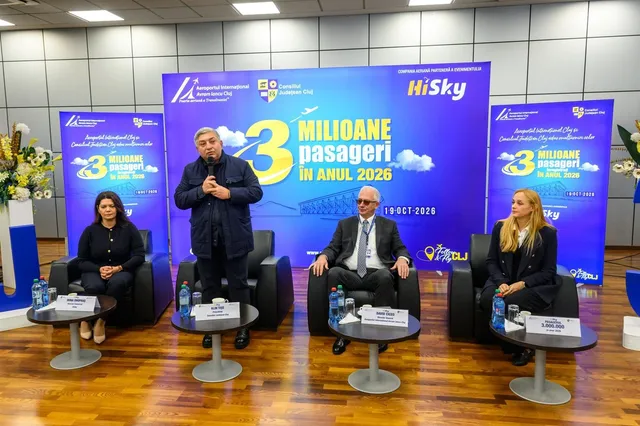 Patru persoane stau în fața unor panouri albastre cu text alb și galben; una ține un microfon, iar în față sunt mese mici.