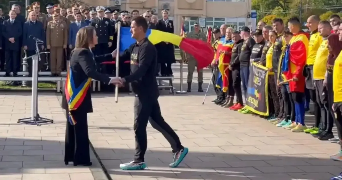 O femeie cu eșarfă tricoloră și un bărbat în haine închise la culoare își strâng mâinile lângă un baston; în spate stau grupuri de oameni și steaguri.