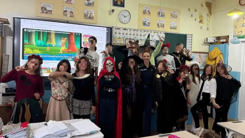 Un grup de persoane costumate stă într-o sală cu bănci, hârtii, afișe și un ecran colorat.