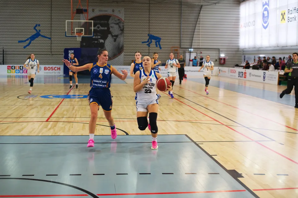 Pe un teren de baschet, o jucătoare în echipament alb ține mingea, iar o jucătoare în echipament albastru aleargă lângă ea.