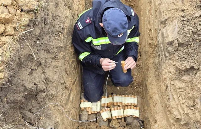 O persoană în uniformă închisă la culoare și șapcă stă ghemuită într-o groapă, lângă mai multe obiecte cilindrice grupate, ținând fire subțiri.