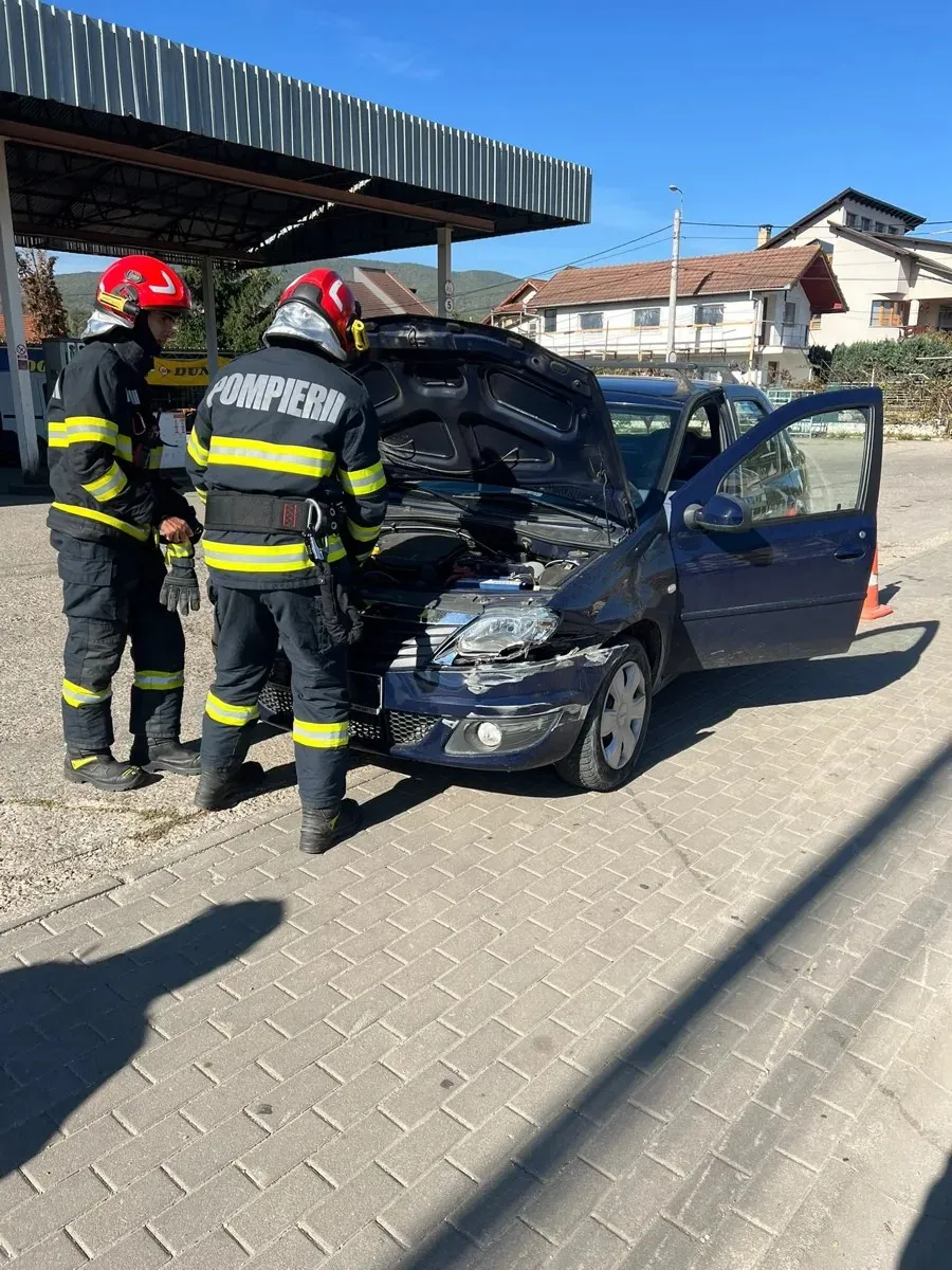 Doi pompieri în uniforme reflectorizante stau lângă un autoturism avariat, cu capota și portiera din dreapta deschise.