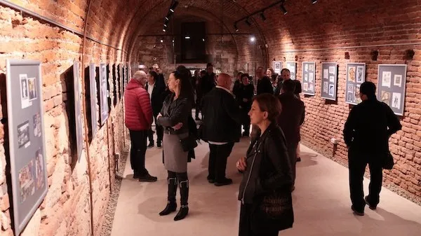 Mai multe persoane stau într-o sală cu pereți de cărămidă și privesc panouri cu imagini expuse de-a lungul pereților.