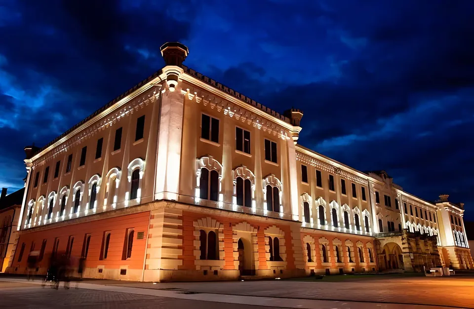 O clădire mare, iluminată, cu fațadă bej, ferestre arcuite și coloane albe, se află lângă o stradă, sub un cer albastru închis.