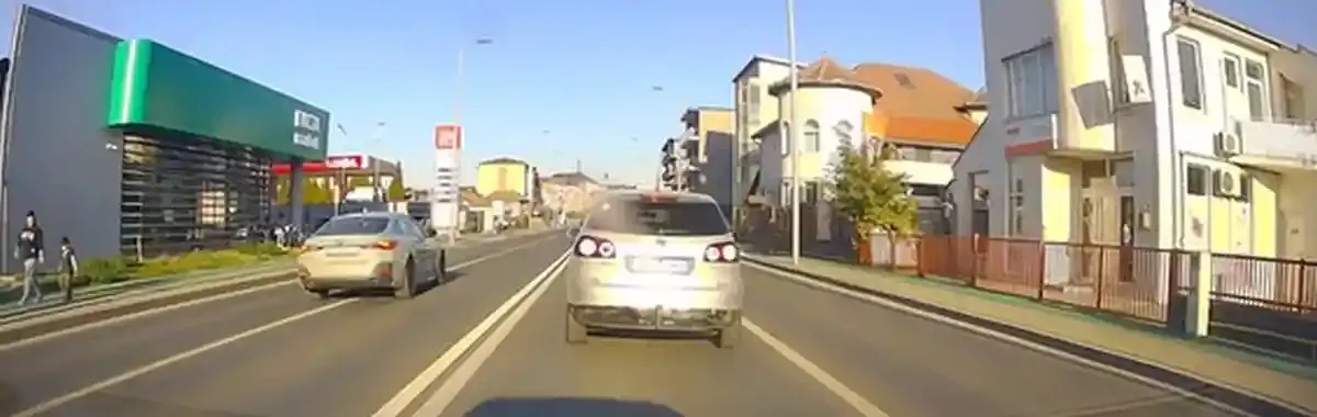 Un autoturism argintiu se află pe o șosea cu marcaje longitudinale, iar de o parte și de alta sunt clădiri, copaci și alte vehicule.