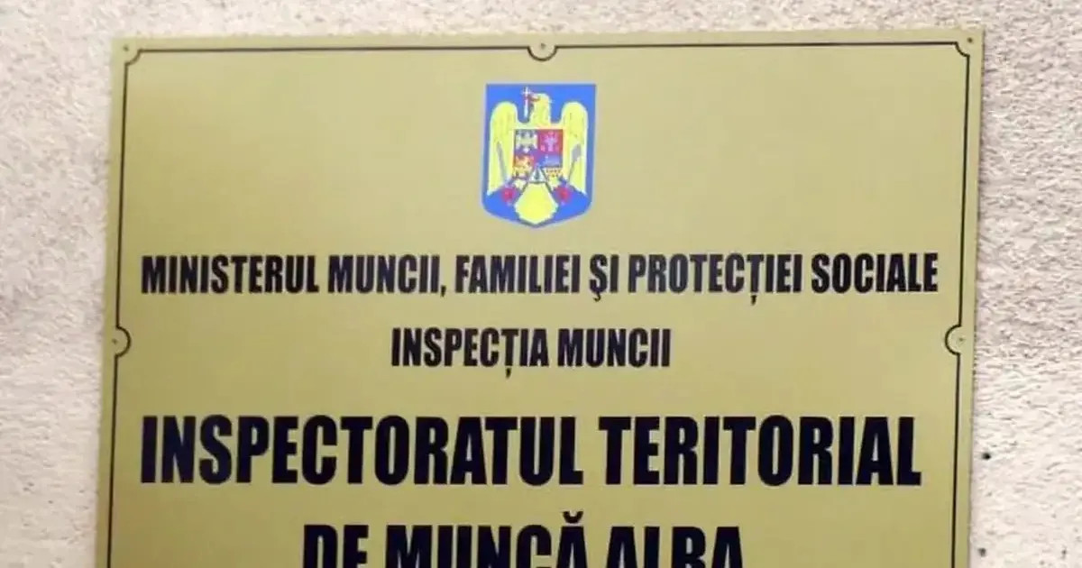 O plăcuță dreptunghiulară galbenă, prinsă pe un perete deschis la culoare, poartă o stemă și inscripții negre despre Inspecția Muncii și ITM Alba.
