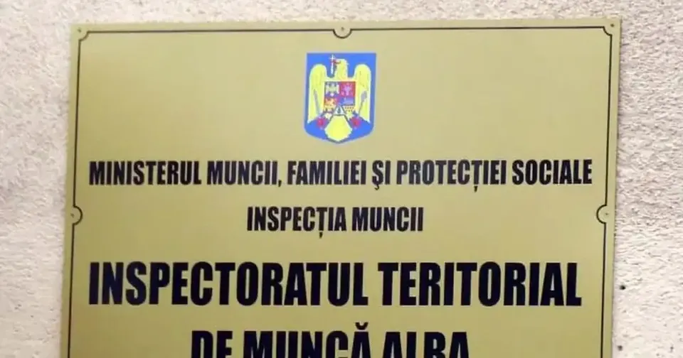 O plăcuță dreptunghiulară galbenă, prinsă pe un perete deschis la culoare, poartă o stemă și inscripții negre despre Inspecția Muncii și ITM Alba.