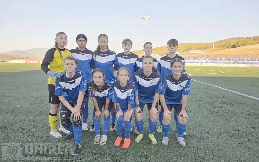 Unsprezece jucătoare de fotbal în echipamente albastre și galbene sunt grupate pe un teren sintetic, cu tribune și dealuri în fundal.