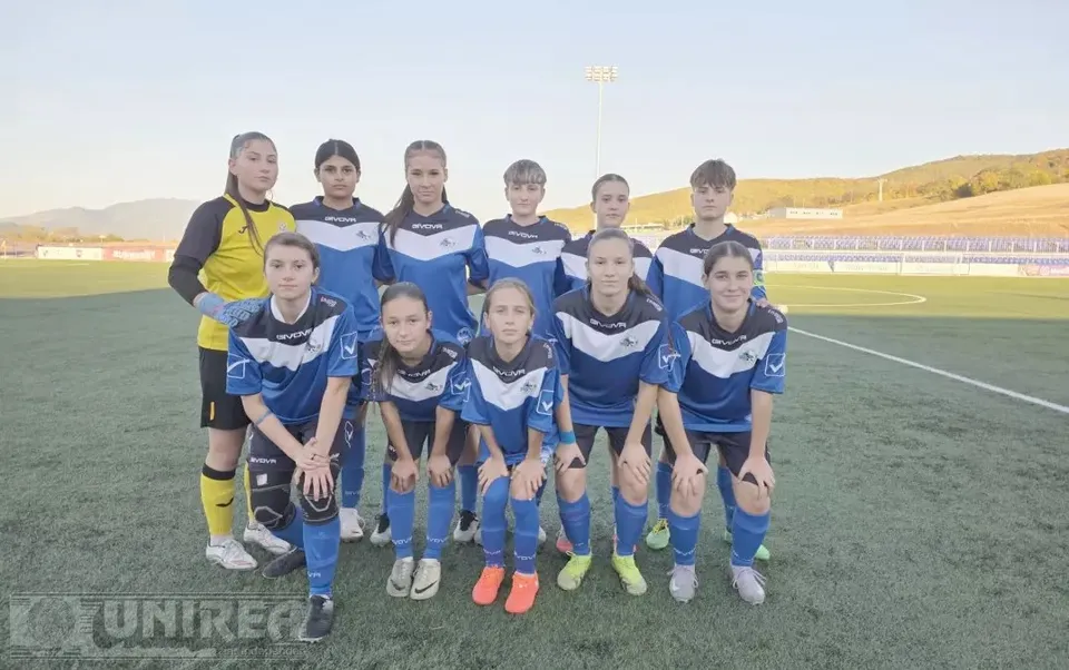 Unsprezece jucătoare de fotbal în echipamente albastre și galbene sunt grupate pe un teren sintetic, cu tribune și dealuri în fundal.