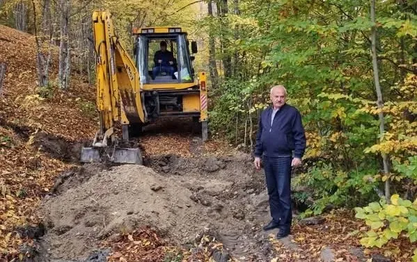 Un excavator galben se află pe un drum de pământ săpat, între copaci; un bărbat în haine închise la culoare stă lângă el.