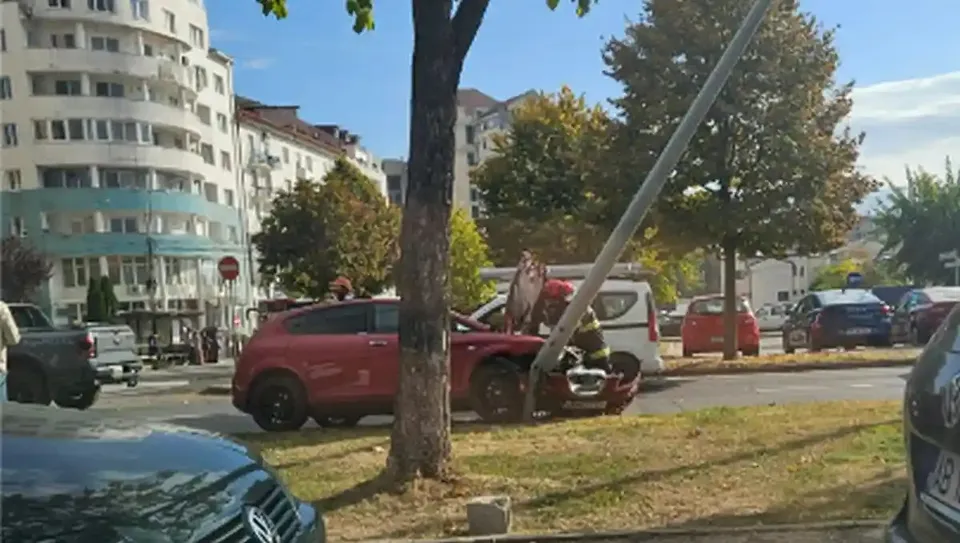 Un SUV roșu cu partea din față avariată este lângă un stâlp înclinat; mai multe vehicule sunt parcate pe marginea drumului.
