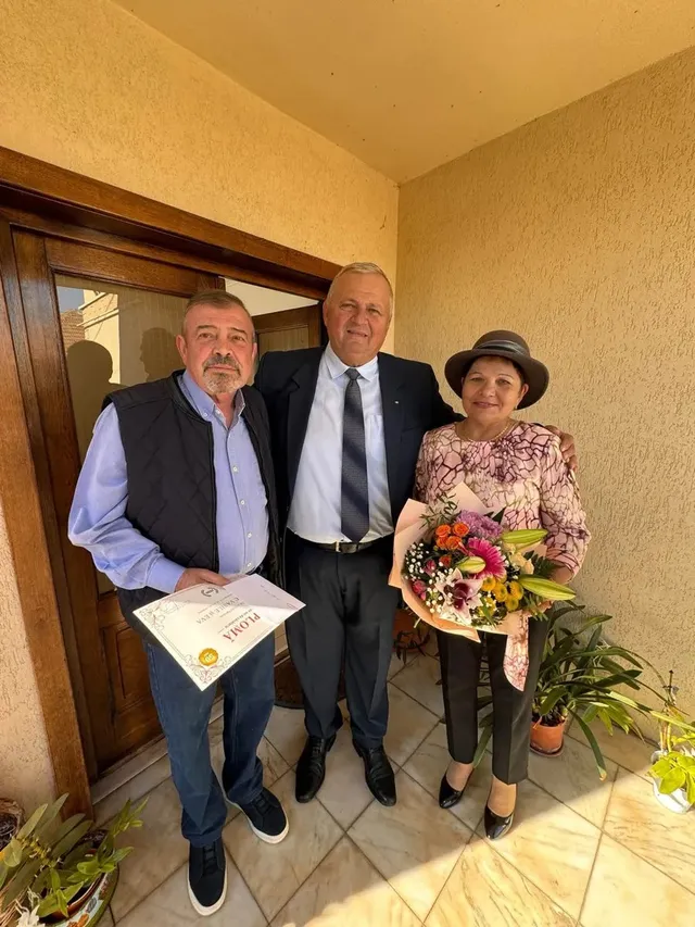 Trei persoane stau într-o încăpere; bărbatul din stânga ține o diplomă, iar femeia din dreapta ține un buchet de flori.
