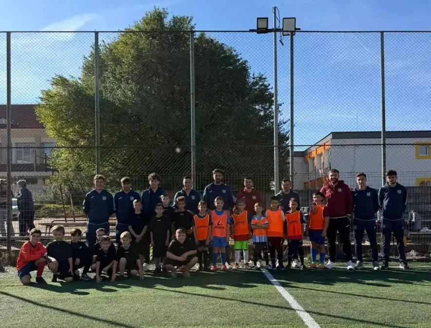 Un grup de adulți și copii, unii în echipament portocaliu, stă pe un teren sintetic, în fața unui gard și a unor clădiri.