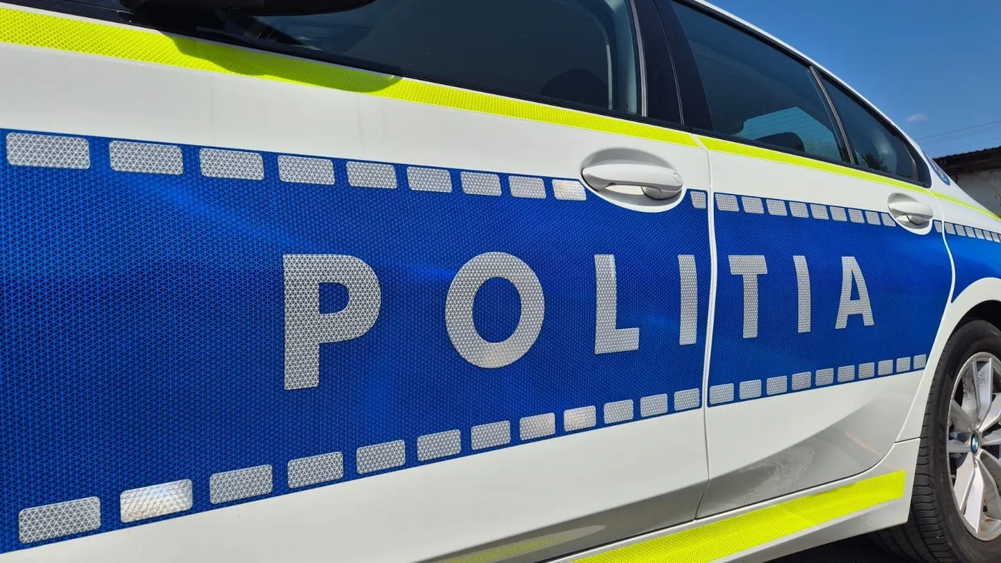 Prim-plan lateral al unui autoturism alb, cu panou albastru inscripționat „POLIȚIA”, dungi galbene reflectorizante, geamuri închise la culoare și o roată.