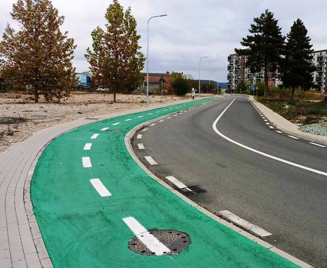 Un drum asfaltat curbat are marcaje albe, o bandă verde, trotuare, stâlpi de iluminat, copaci și clădiri în fundal.