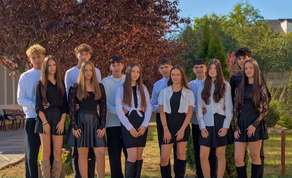 Un grup de tineri stă în aer liber, în fața unor copaci, purtând ținute predominant negre și albe.