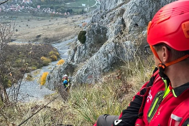 În prim-plan, o persoană cu cască roșie și jachetă roșie stă lângă o stâncă; mai jos se vede o persoană cu cască.