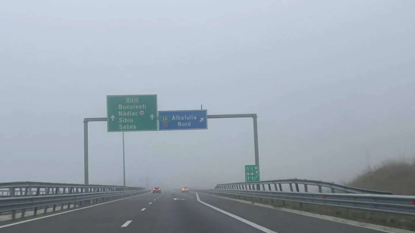 O șosea cu parapete metalice se pierde în ceață; deasupra ei sunt indicatoare rutiere, iar în depărtare se văd două vehicule.