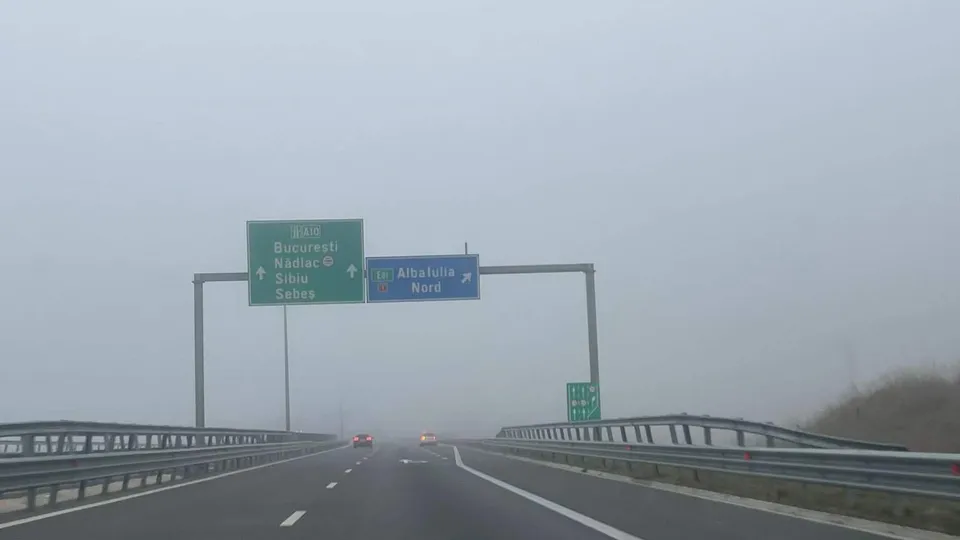 O șosea cu parapete metalice se pierde în ceață; deasupra ei sunt indicatoare rutiere, iar în depărtare se văd două vehicule.