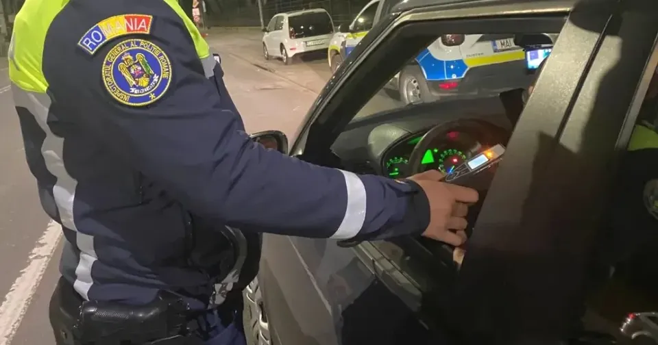 O persoană în uniformă albastră cu elemente reflectorizante ține mâna la fereastra unui autoturism negru, lângă un dispozitiv mic.