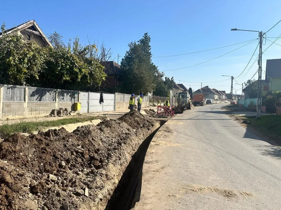 Un șanț cu pământ excavat se întinde de-a lungul unei străzi; în depărtare se văd câteva persoane și un utilaj.