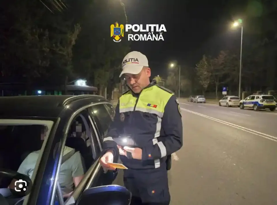 Un polițist în uniformă reflectorizantă stă lângă o mașină, iar o persoană se află la volan; pe șosea se văd alte vehicule și lămpi.
