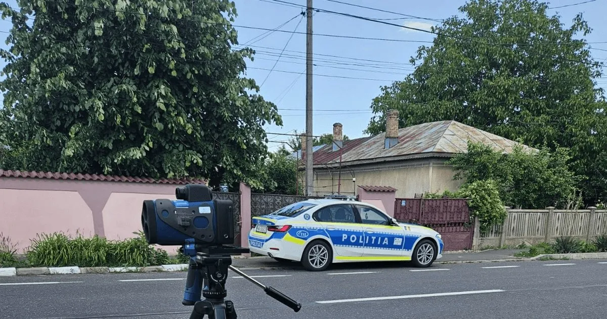 Un dispozitiv negru este montat pe un trepied lângă o mașină albă și albastră inscripționată „POLIȚIA”, pe o stradă.