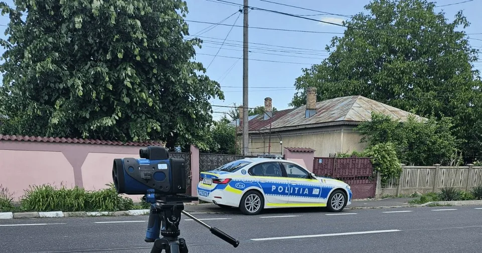 Un dispozitiv negru este montat pe un trepied lângă o mașină albă și albastră inscripționată „POLIȚIA”, pe o stradă.