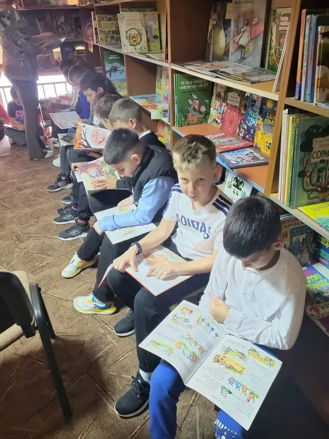 Mai mulți copii stau pe scaune și privesc cărți deschise, lângă rafturi pline cu cărți; în prim-plan, un copil ține o carte ilustrată.