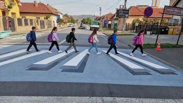 Mai multe persoane cu rucsacuri traversează o trecere cu marcaje albe și gri, în timp ce mașini și clădiri se văd în fundal.