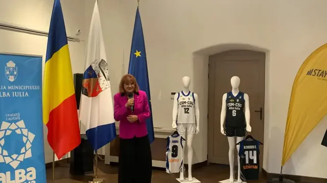 O femeie în sacou roz ține un microfon lângă două manechine îmbrăcate în echipamente de baschet, tricouri expuse și mai multe steaguri.