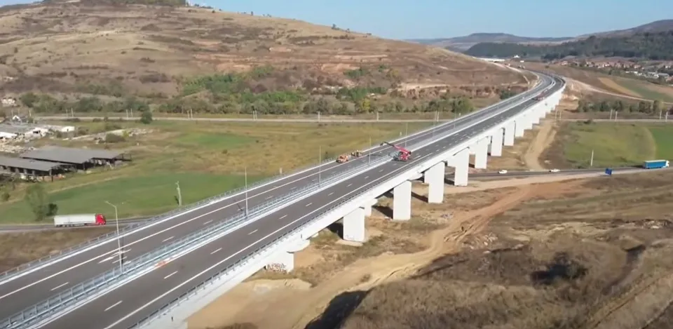 Un viaduct rutier lung, cu două căi asfaltate și piloni albi, traversează un teren deluros; câteva vehicule se află pe șosea.