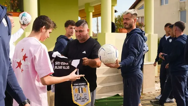 Mai mulți bărbați stau lângă o clădire; unul ține o minge albă, iar alții țin un tricou negru și un fanion.