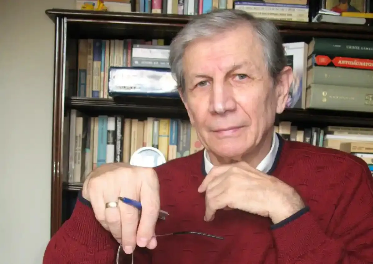 Un bărbat cu păr cărunt, îmbrăcat într-un pulover roșu, stă în fața unei biblioteci, ținând ochelari și purtând un inel.