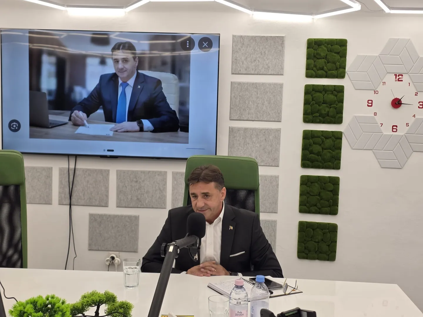 Un bărbat în sacou închis stă la o masă albă, lângă un microfon; pe ecranul din spate apare un alt bărbat în costum.