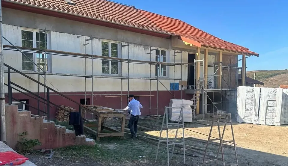 O clădire cu acoperiș din țiglă roșie are schele în față, iar două persoane se află lângă materiale și echipamente de construcție.