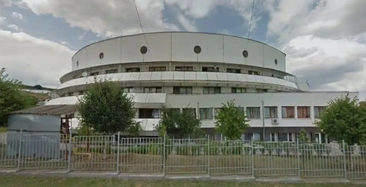 O clădire albă cu mai multe niveluri, ferestre dreptunghiulare și circulare se află în spatele unui gard metalic, între copaci, sub un cer înnorat.