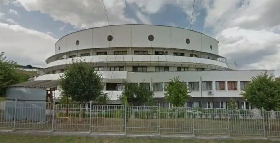 O clădire albă cu mai multe niveluri, ferestre dreptunghiulare și circulare se află în spatele unui gard metalic, între copaci, sub un cer înnorat.