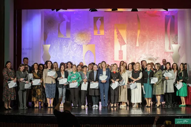 Un grup numeros de adulți și tineri stă pe o scenă, majoritatea ținând diplome, iar în prim-plan apare silueta unui fotograf.