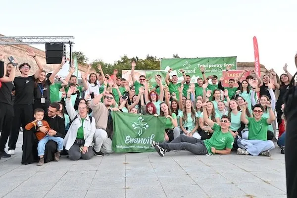Un grup numeros de adulți și copii, îmbrăcați predominant în verde, pozează cu brațele ridicate în spatele unui banner verde.