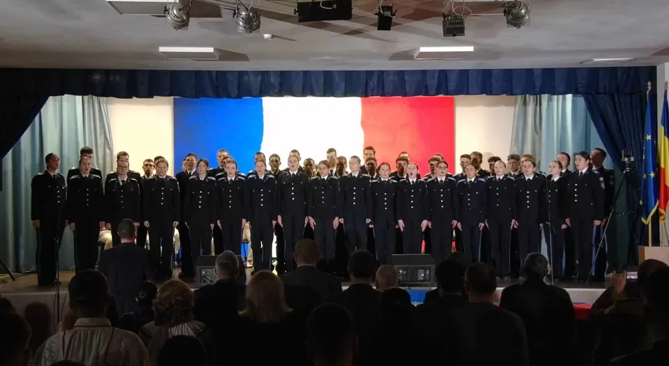 Un grup de persoane în uniforme închise la culoare stă pe scenă, în fața unui fundal tricolor, iar în prim-plan se văd spectatori.