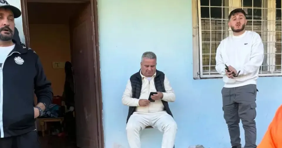Trei bărbați sunt lângă un perete albastru: unul stă pe scaun cu un telefon, altul stă în picioare lângă fereastră.