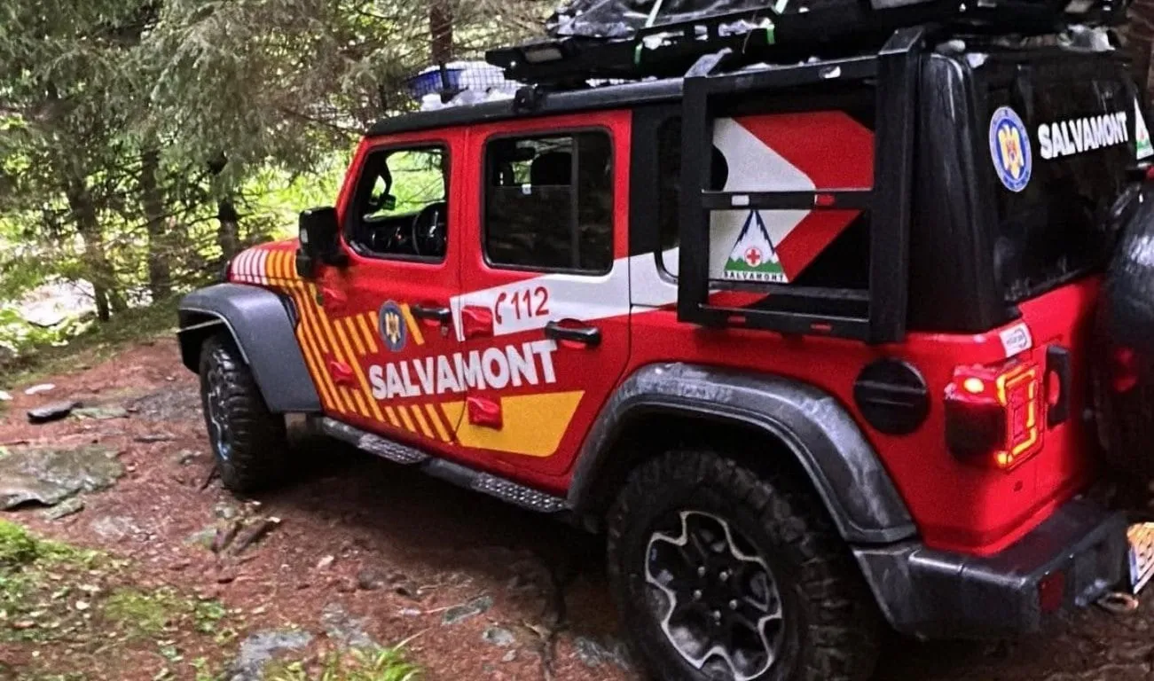 Un vehicul de teren roșu și negru, inscripționat „SALVAMONT” și „112”, este parcat pe o potecă împădurită.