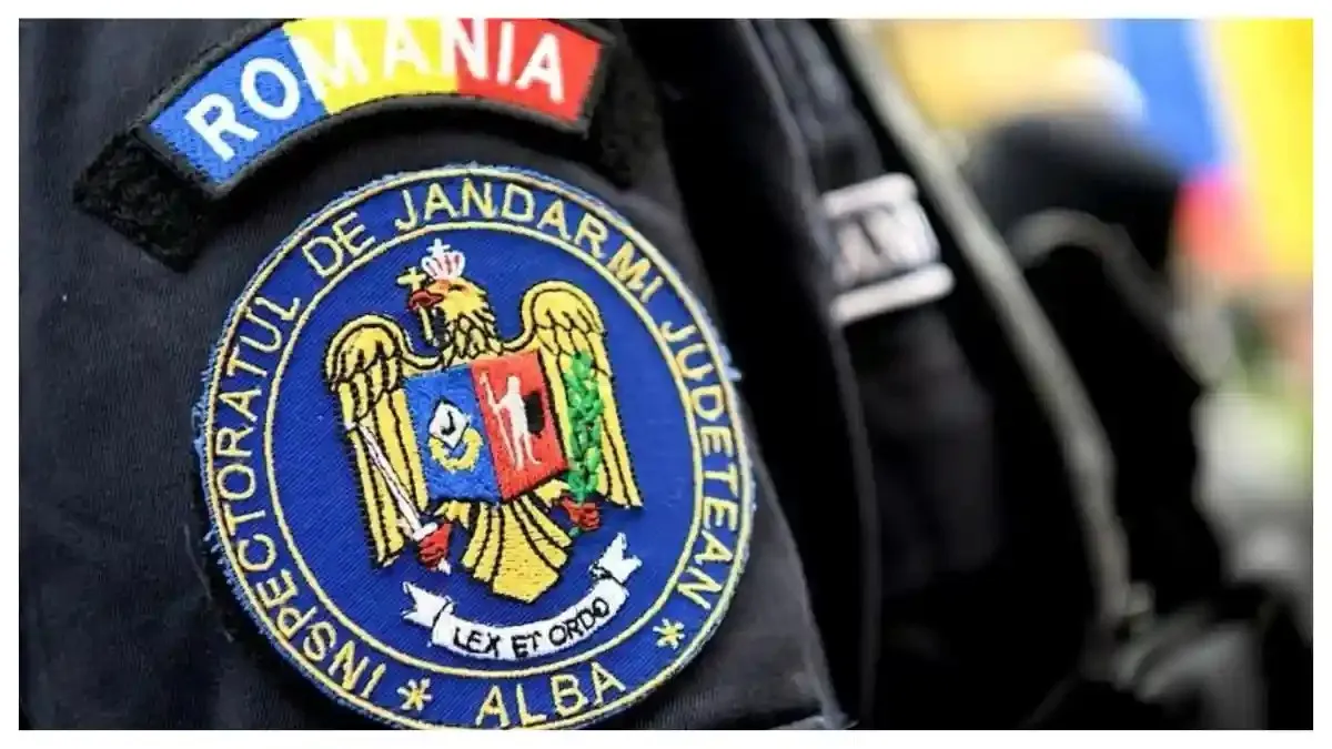Prim-plan al unei uniforme negre cu ecusoane brodate „ROMÂNIA” și „INSPECTORATUL DE JANDARMI JUDEȚEAN ALBA”, lângă o a doua uniformă neclară.
