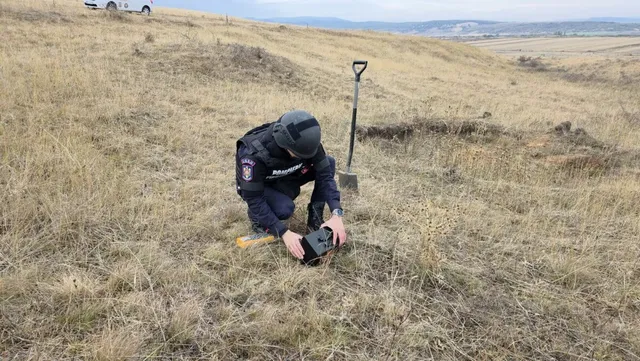 O persoană în echipament închis la culoare stă ghemuită pe un câmp cu iarbă uscată, lângă un obiect negru și o lopată.