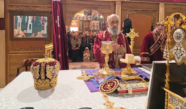 Doi clerici în veșminte vișinii stau în spatele unei mese cu vase aurii, cărți și o coroană decorată; mai multe persoane se află în fundal.