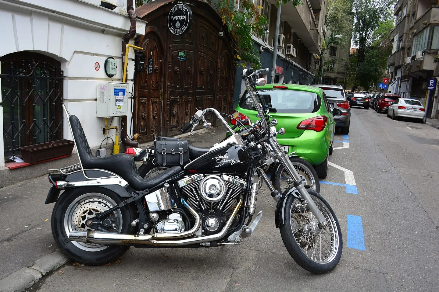 O motocicletă neagră cu elemente cromate este parcată lângă o mașină verde, pe o stradă mărginită de clădiri.